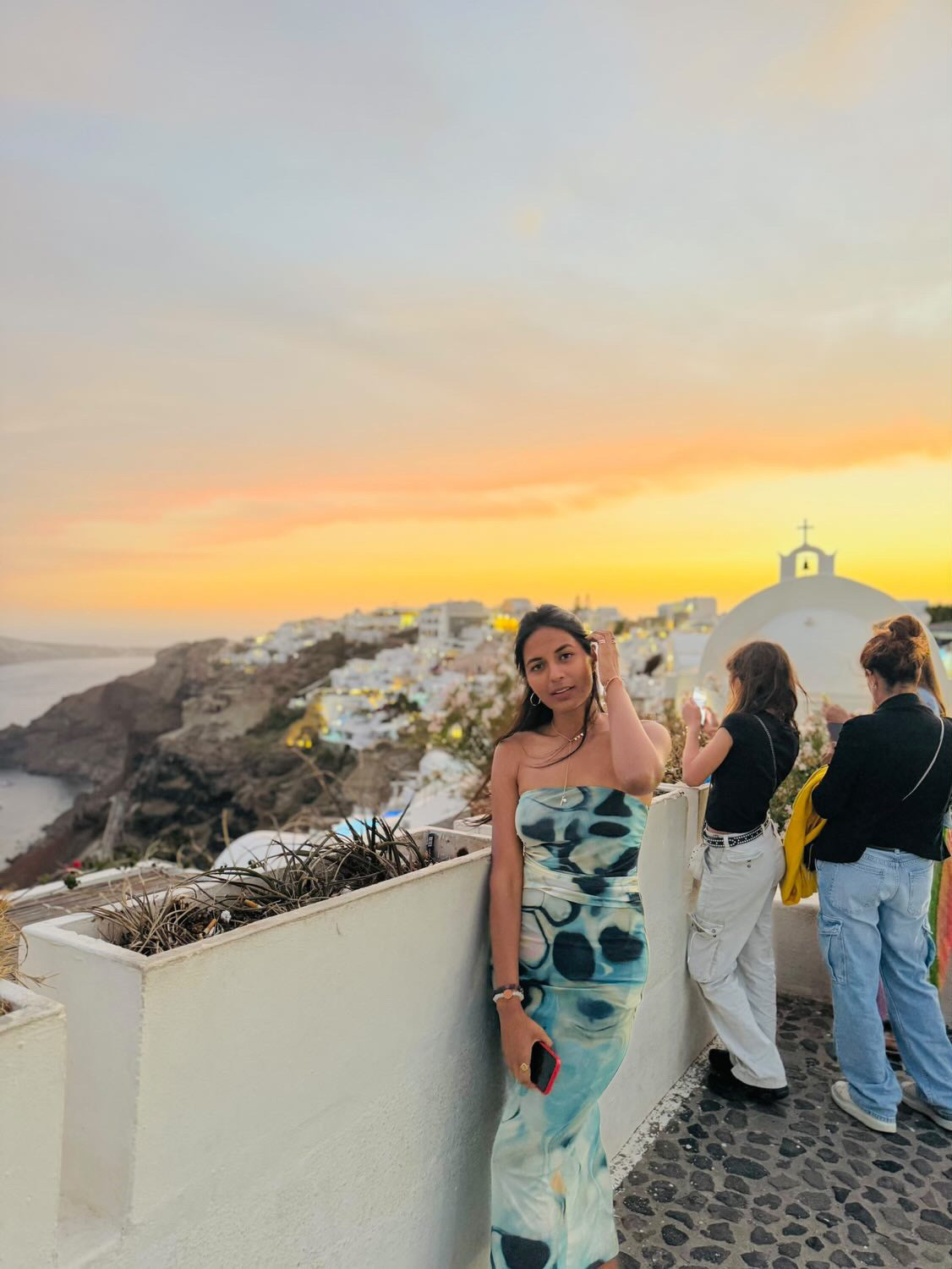 Strapless beige patch-dye tube dress, calf-length, form-fitting, worn by a woman along a railing at sunset in Santorini.