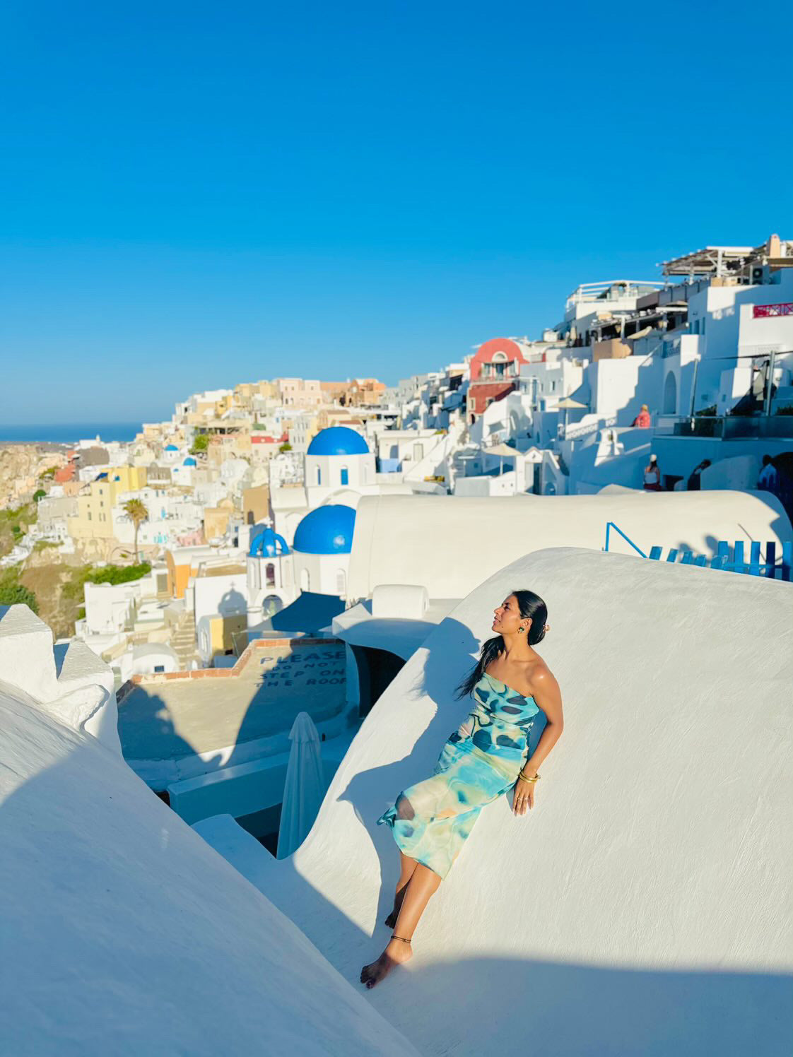 Beige patch-print tie-dye tulle tube dress worn by a woman seated on white Santorini walls with blue domes.