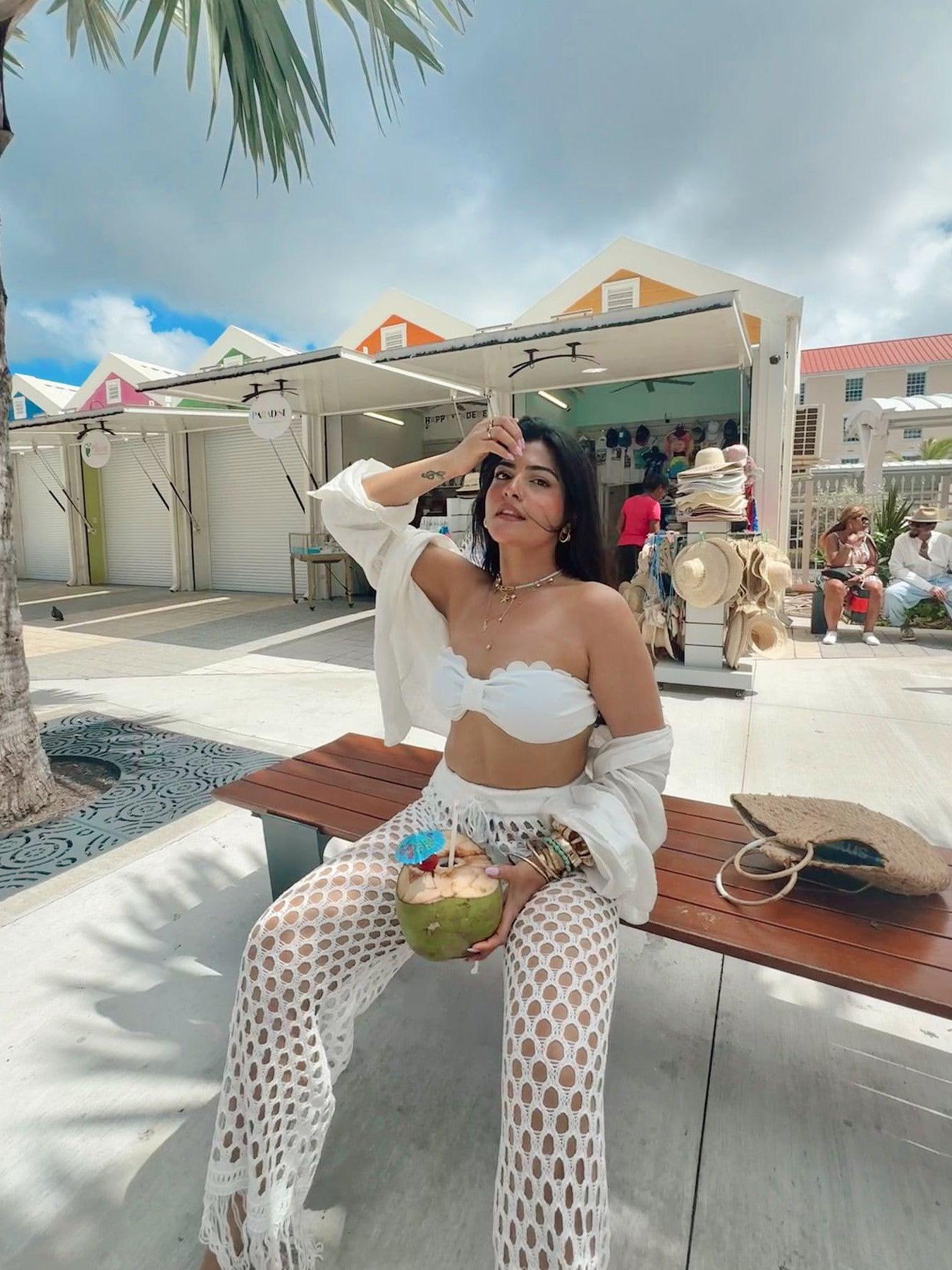 Woman wearing white scalloped-edge crochet bralette and matching mesh crochet pants, seated on a bench with a coconut drink.