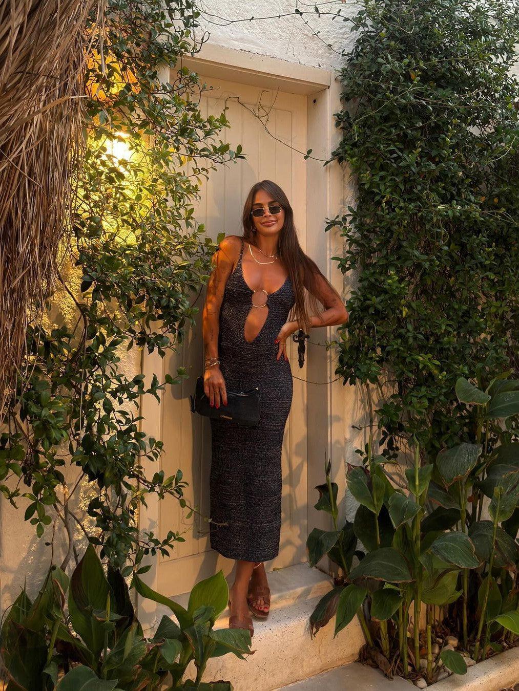 Dark knit backless spaghetti-strap midi dress with a front ring cutout, worn by a woman beside a door among greenery.