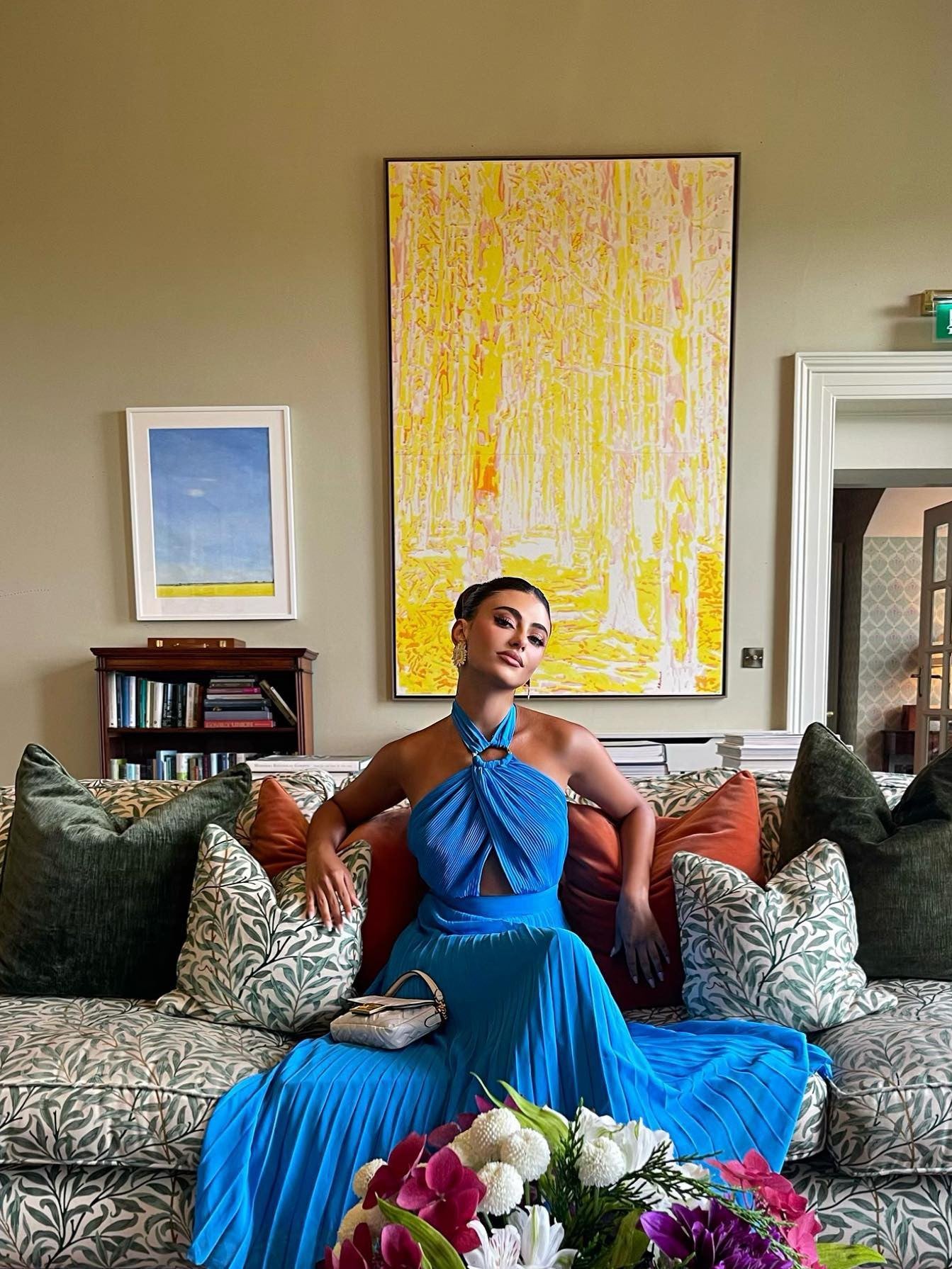 Blue halter-neck pleated dress with cutout and buckle on a woman seated on a patterned sofa; clutch on lap.