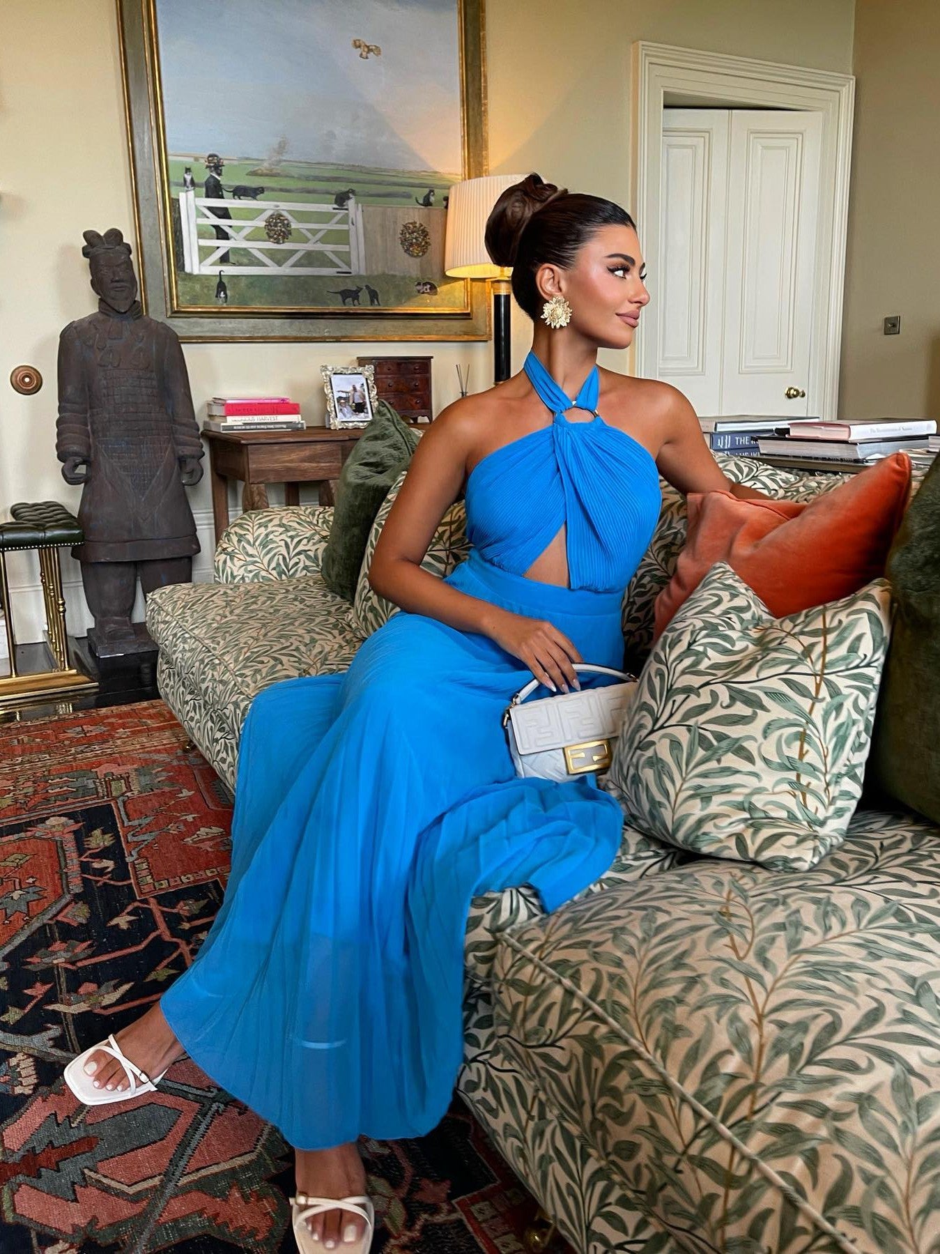Bright blue halter-neck pleated dress with waist cutout and flowing skirt; white sandals, white purse, and floral earrings.
