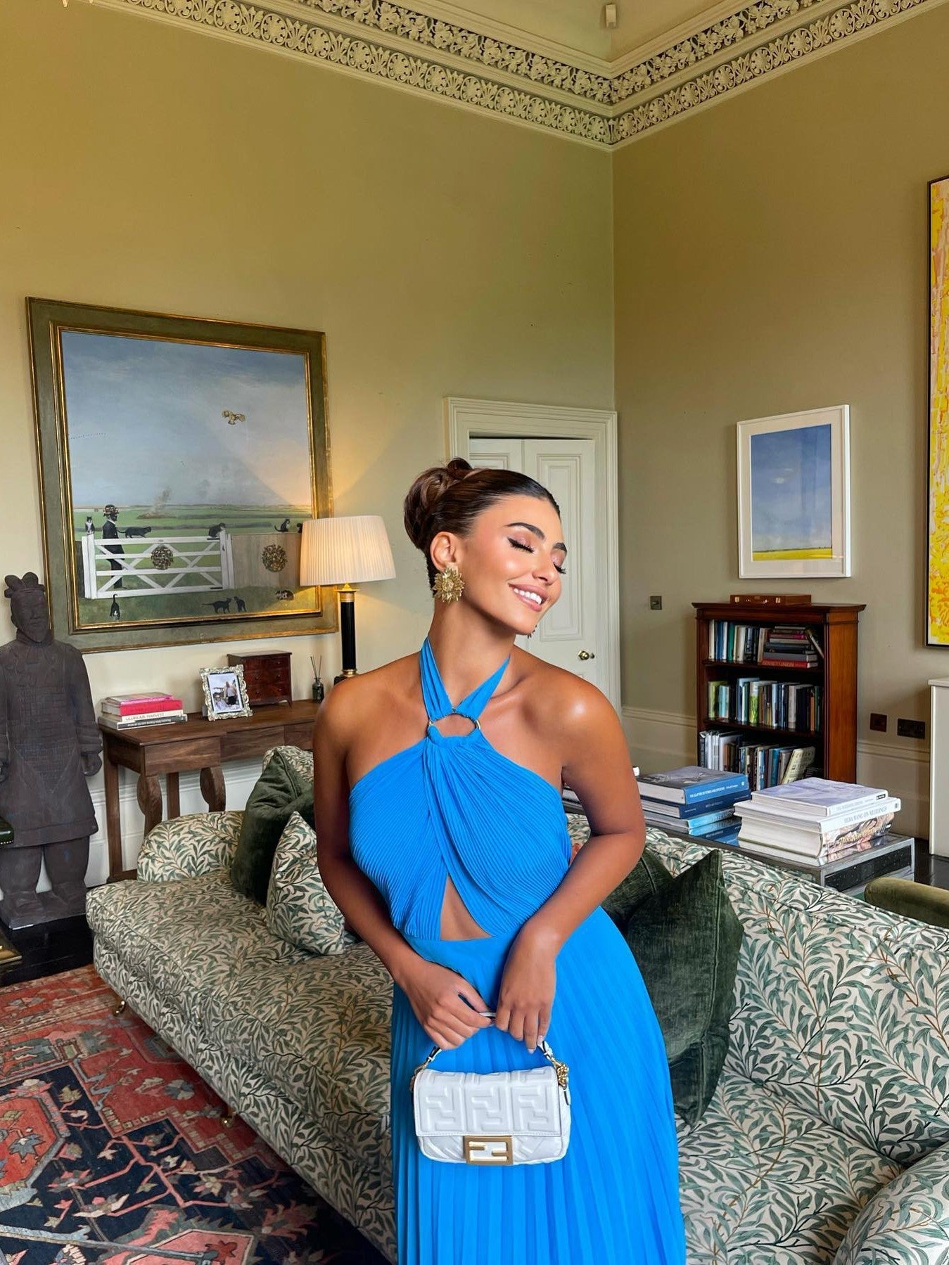 Cobalt-blue halterneck cutout pleated dress with buckle on a woman in a living room, holding a white quilted handbag.
