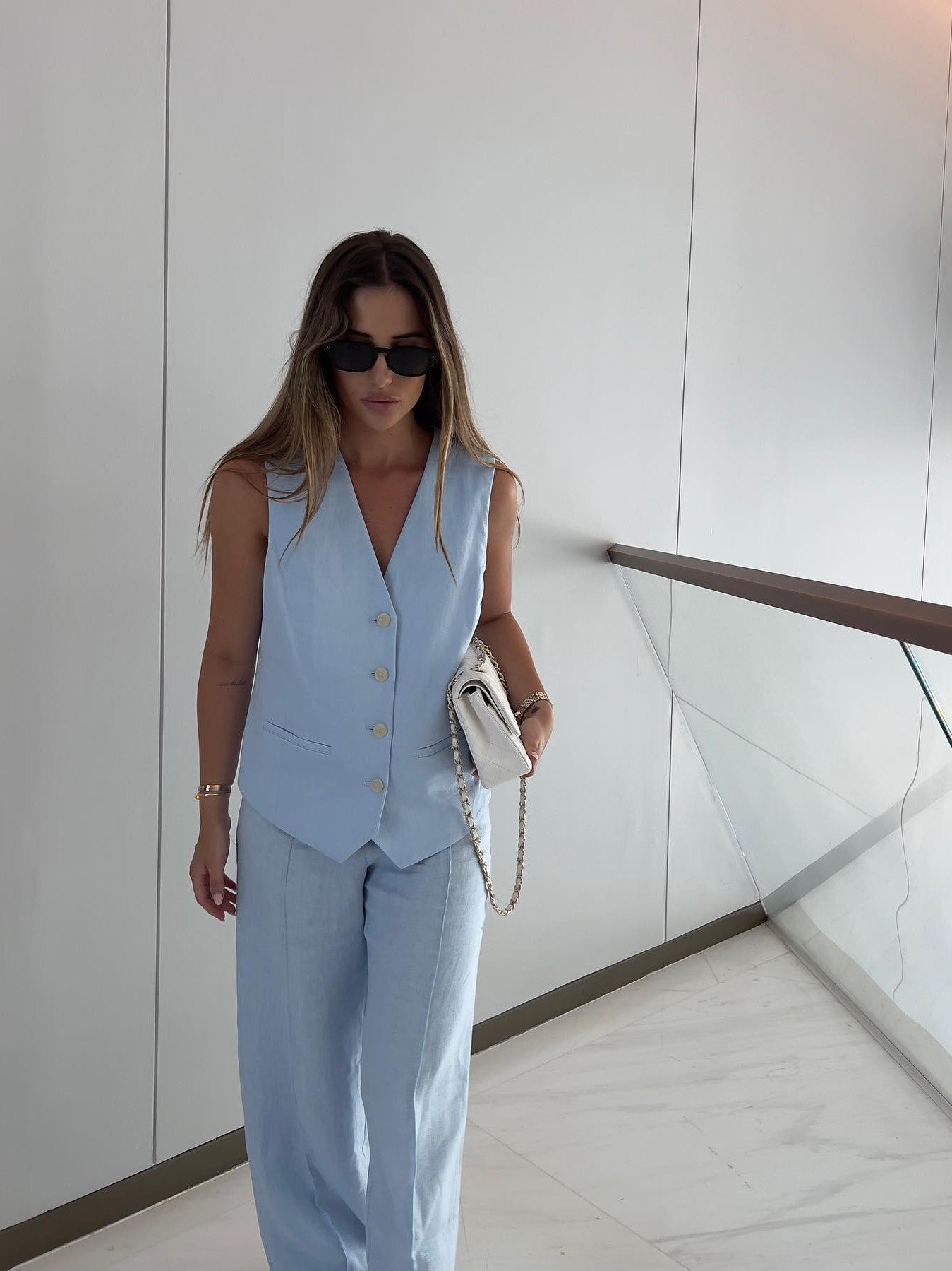 Light-blue waistcoat with beige buttons over wide-leg pants; woman in sunglasses holding a white bag with chain strap.