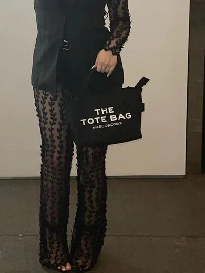 Person wearing black floral mesh sheer flared pants, holding a black tote bag that reads THE TOTE BAG MARC JACOBS.