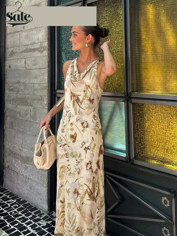 Woman in cream floral cowl-neck maxi dress with beige handbag and gold earrings, standing by colorful glass windows.