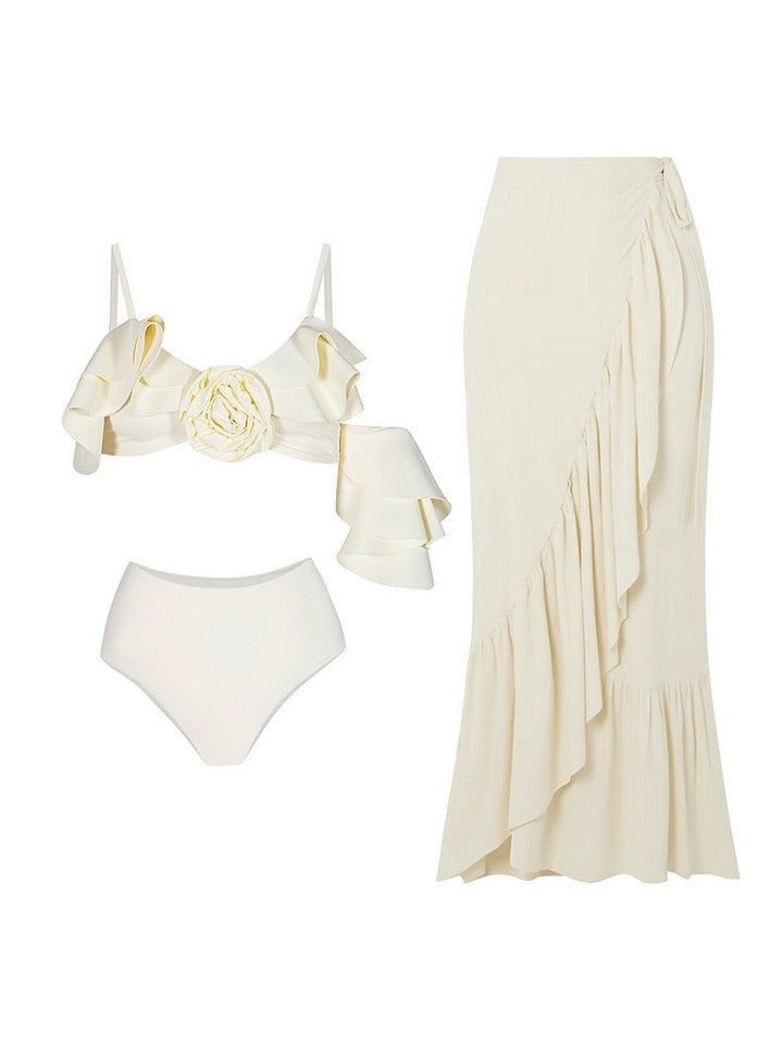 Cream three-piece swimsuit: ruffled floral-front bikini top, high-waisted bottoms, and a long asymmetric-ruffle wrap skirt.