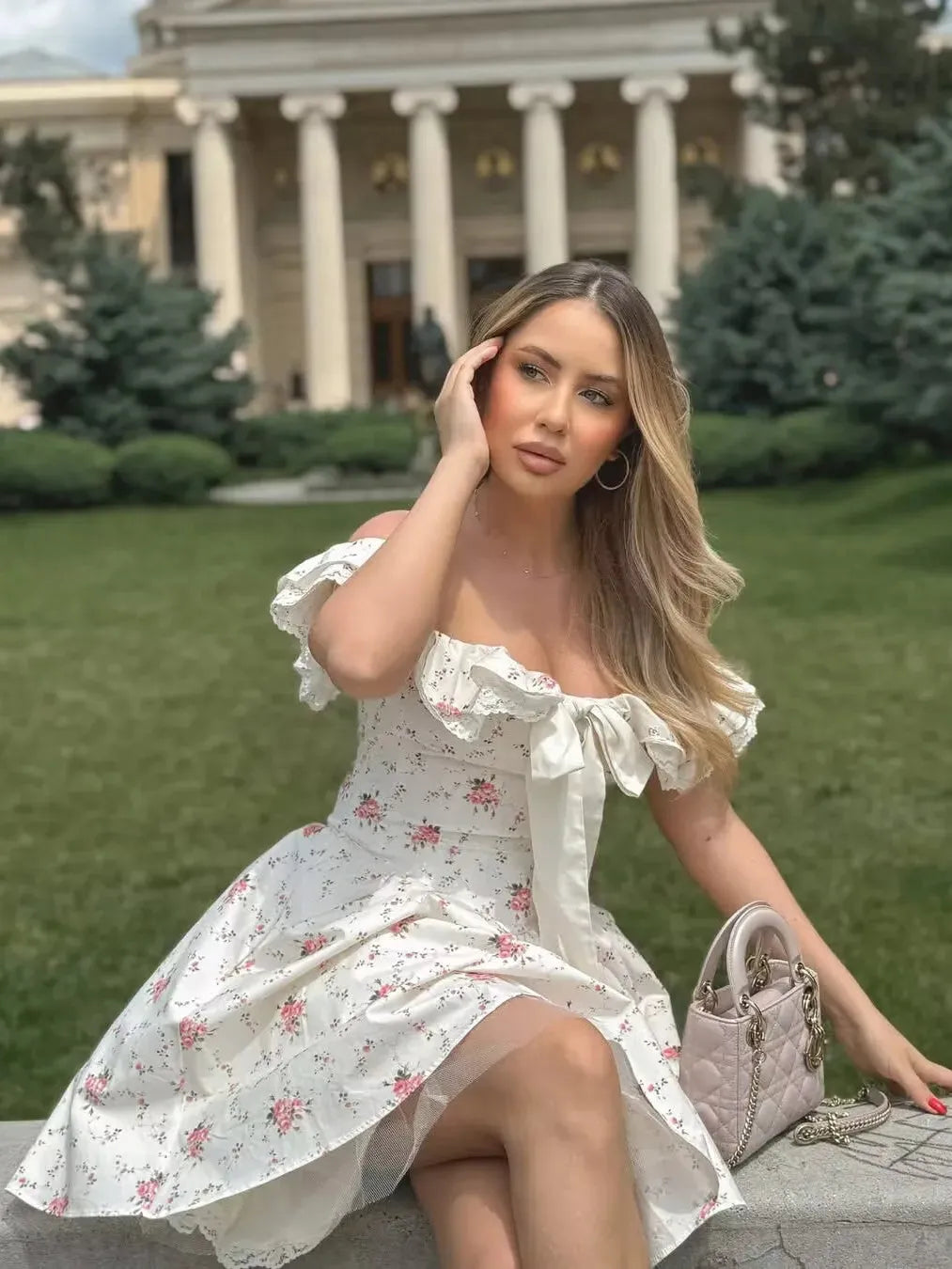 White off-shoulder mini dress with pink rose print, ruffled neckline and front bow, outdoors with a blush quilted handbag.