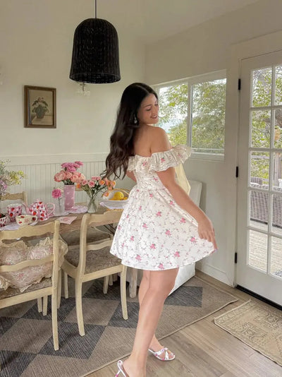 Woman with long dark hair in a white floral off-shoulder mini dress, posing in a bright dining room with pink flowers on the table.