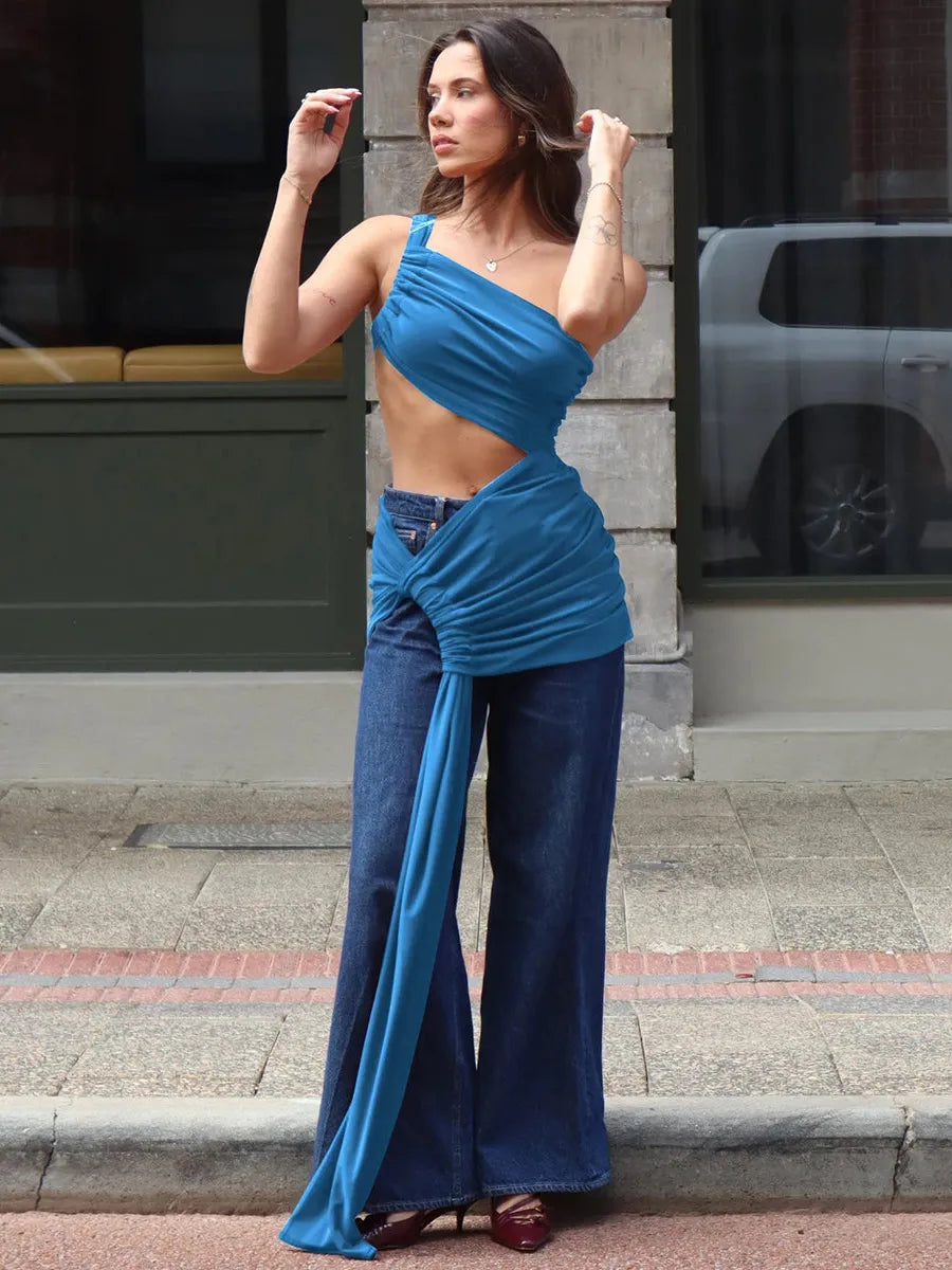 Woman in blue one-shoulder crop top with cut-out and a long draped sash tied at the hip over wide-leg jeans, posing on a city sidewalk.