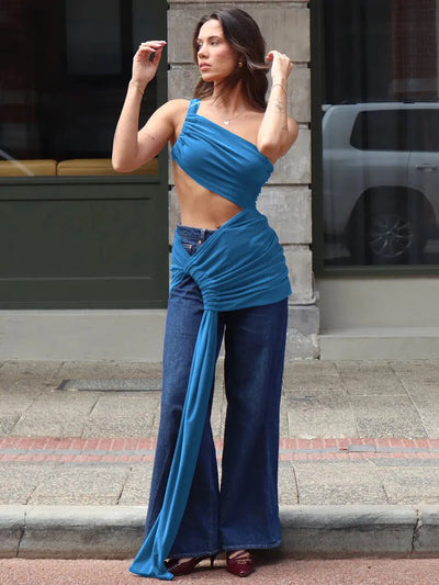 Woman in blue one-shoulder crop top with cut-out and a long draped sash tied at the hip over wide-leg jeans, posing on a city sidewalk.