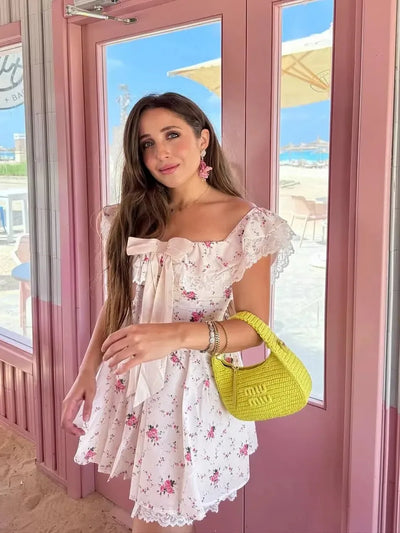 Woman in white floral off-shoulder mini dress with lace trim and bow, carrying a bright yellow woven handbag.