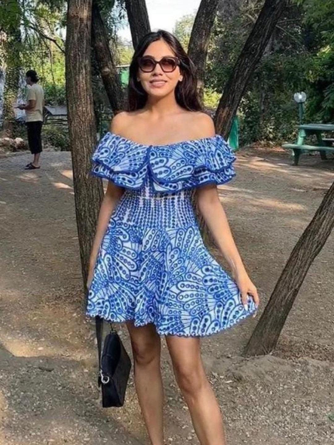 Off-shoulder blue patterned short dress with a ruffled neckline and smocked waist, worn by a woman in a park.