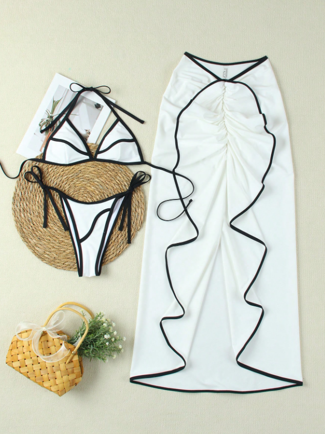 White bikini with black piping and ruffle trim; triangle top with halter ties, side-tie bottoms, paired with a long white ruffle-edged cover-up skirt and straw bag.