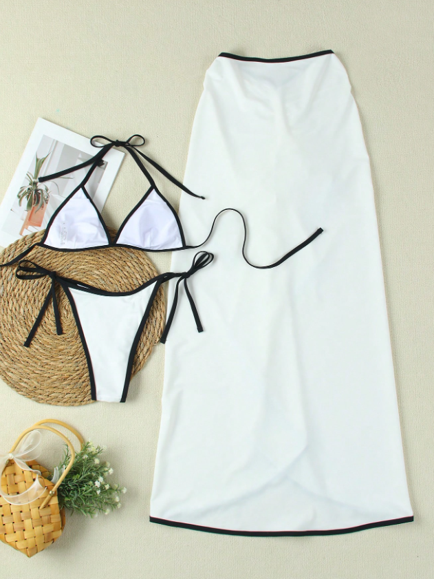 Three-piece white bikini set with black trim: triangle top, tie-side bottoms, and a long white cover-up skirt laid on a beige surface.