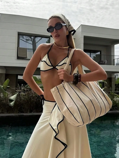 Woman in a white bikini with black piping and ruffle trim, by a pool, holding a striped beige tote and a white ruffle skirt.
