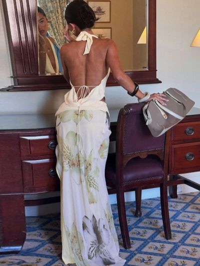 Back view of a woman in a white draped halter top and floral maxi skirt, back tie, beside a burgundy chair with a taupe handbag.