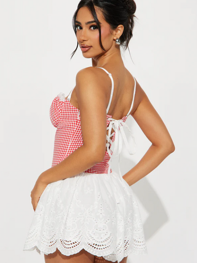 Side view of a woman in a red gingham ruffle top with white lace-up back and a white eyelet mini skirt.