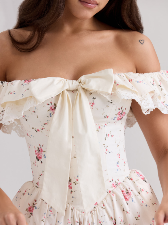 Model wearing off-shoulder ivory floral mini dress with lace trim, cream bow at bodice, ruffled sleeves, pink rose print.