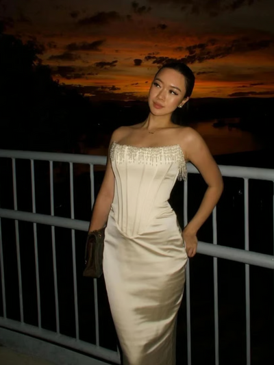 Off-white satin strapless corset gown with pearl fringe along the neckline, posed on a balcony at sunset.