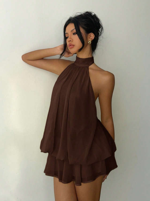 Chocolate brown halter-neck layered mini dress with pleated front panels and two-tier ruffle hem on a model against a light wall.