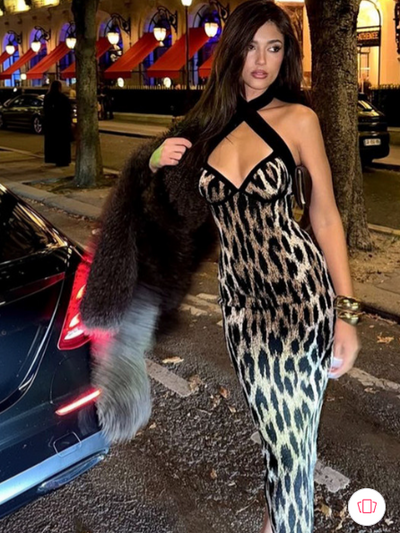 Woman in halter-neck abstract leopard-print maxi dress with cross straps, fur coat on arm, city street at night.