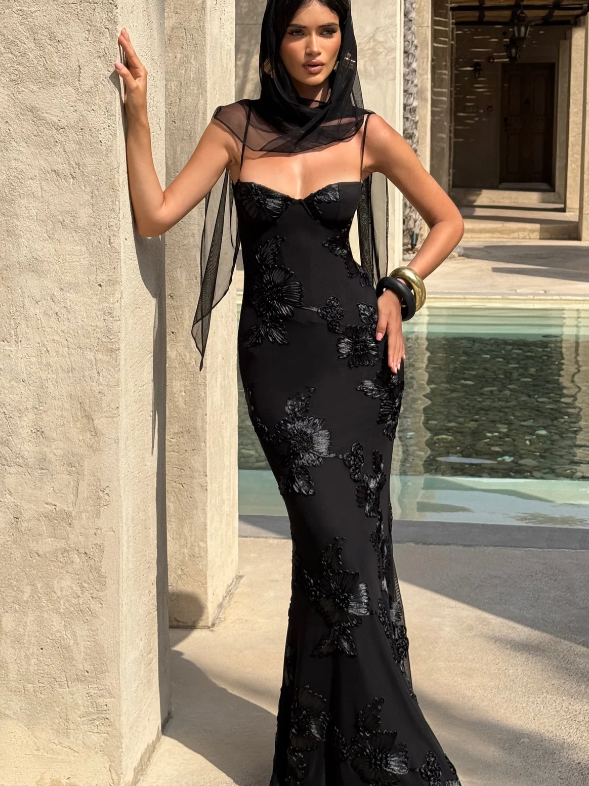 Model in a black floral jacquard corset mermaid maxi dress with sheer shoulder veil, by stone columns and a pool.