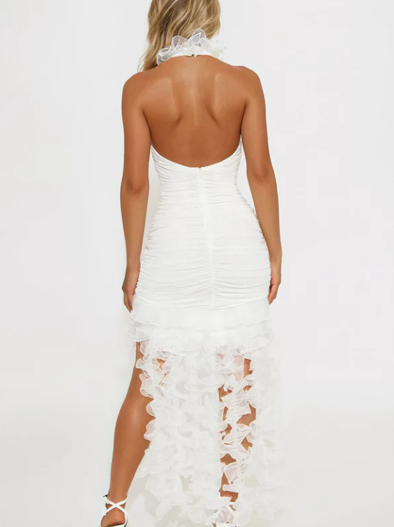 Back view of a white halter mini dress with a ruched bodice and a sheer, petal-embellished high-low skirt, paired with white strappy heels.