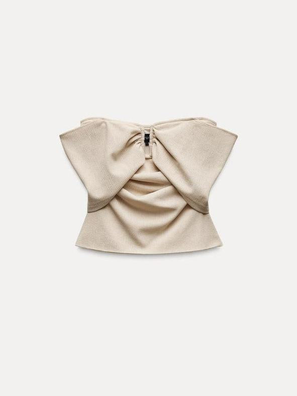 Beige off-shoulder tube top with oversized bow-style flaps across the upper back, draped center panel, and visible back zipper.