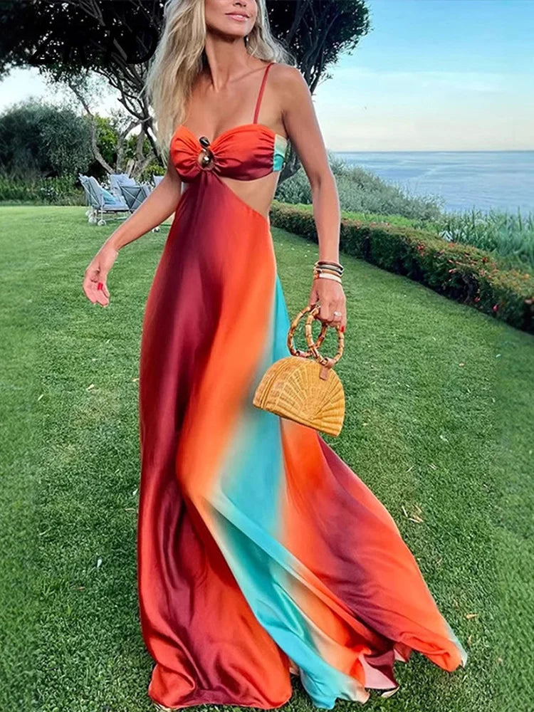Woman in gradient tie-dye cutout maxi with straps; orange-red-teal hues flow down, holding a bamboo handbag on a grassy coastal lawn.