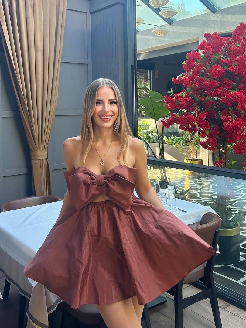 Rust-brown strapless mini dress with a large front bow and full A-line skirt, worn by a smiling woman beside red bougainvillea outdoors.