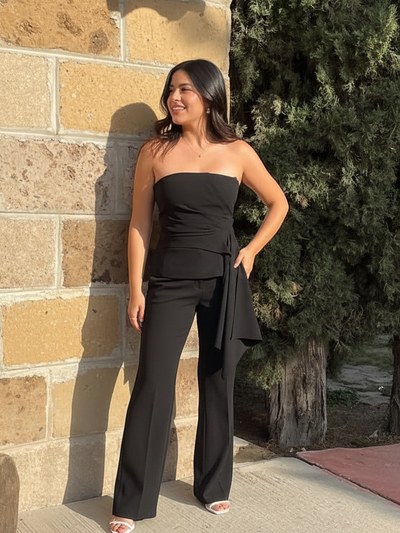 Woman wearing a black asymmetric tube-top with waist wrap and wide-leg pants, posed against a beige stone wall.
