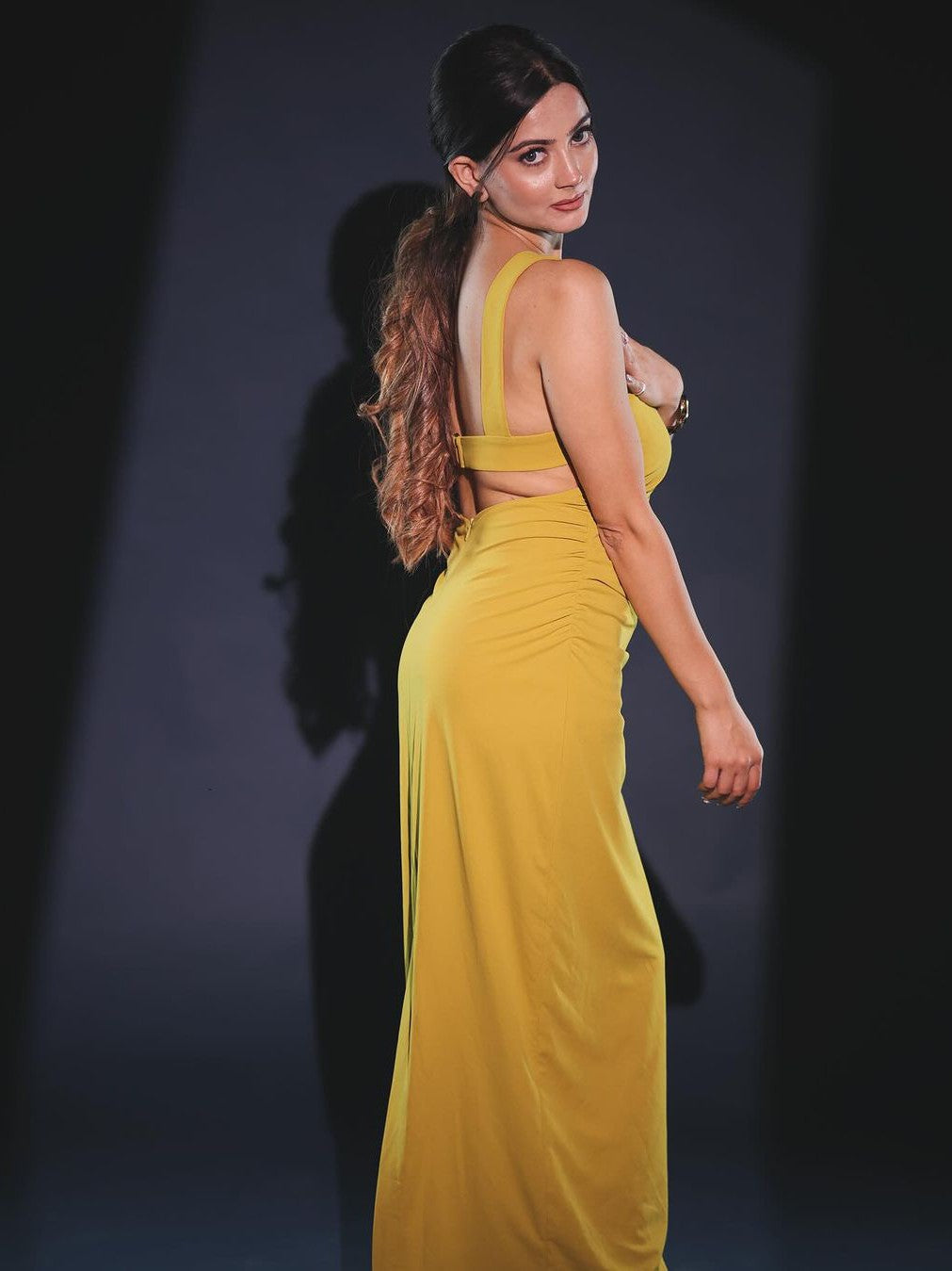 Side view of a woman wearing a yellow backless spaghetti-strap gown with a long flowing skirt against a dark backdrop.