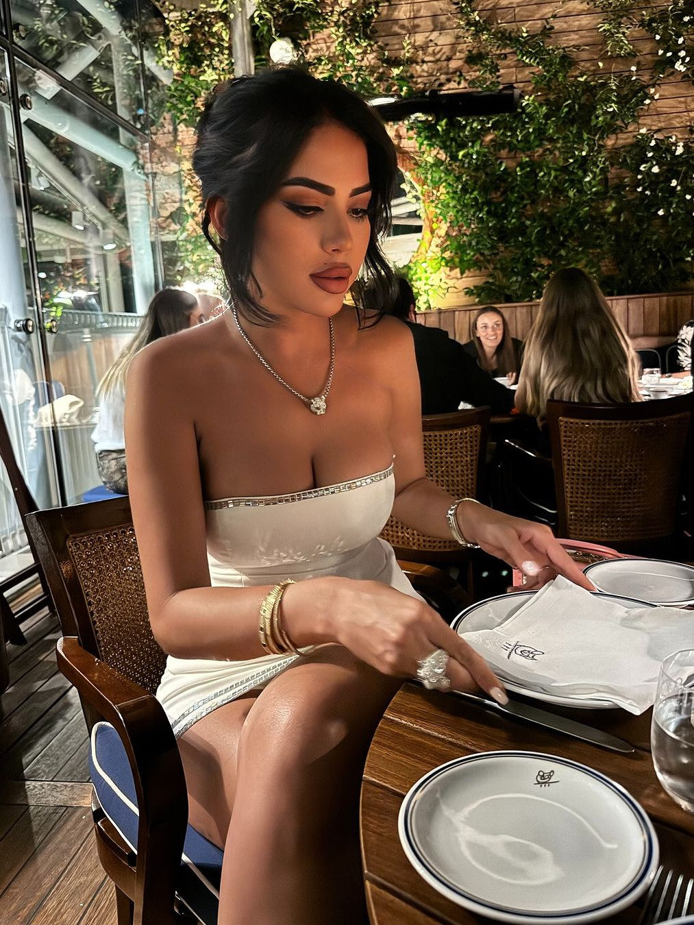 Woman in a white rhinestone-trim tube top and matching skort, seated at a table with a silver necklace and bracelets.