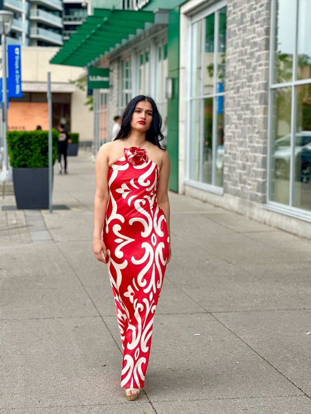 Red halter-backless maxi dress with white floral print and a large neckline flower, worn by woman on city sidewalk.