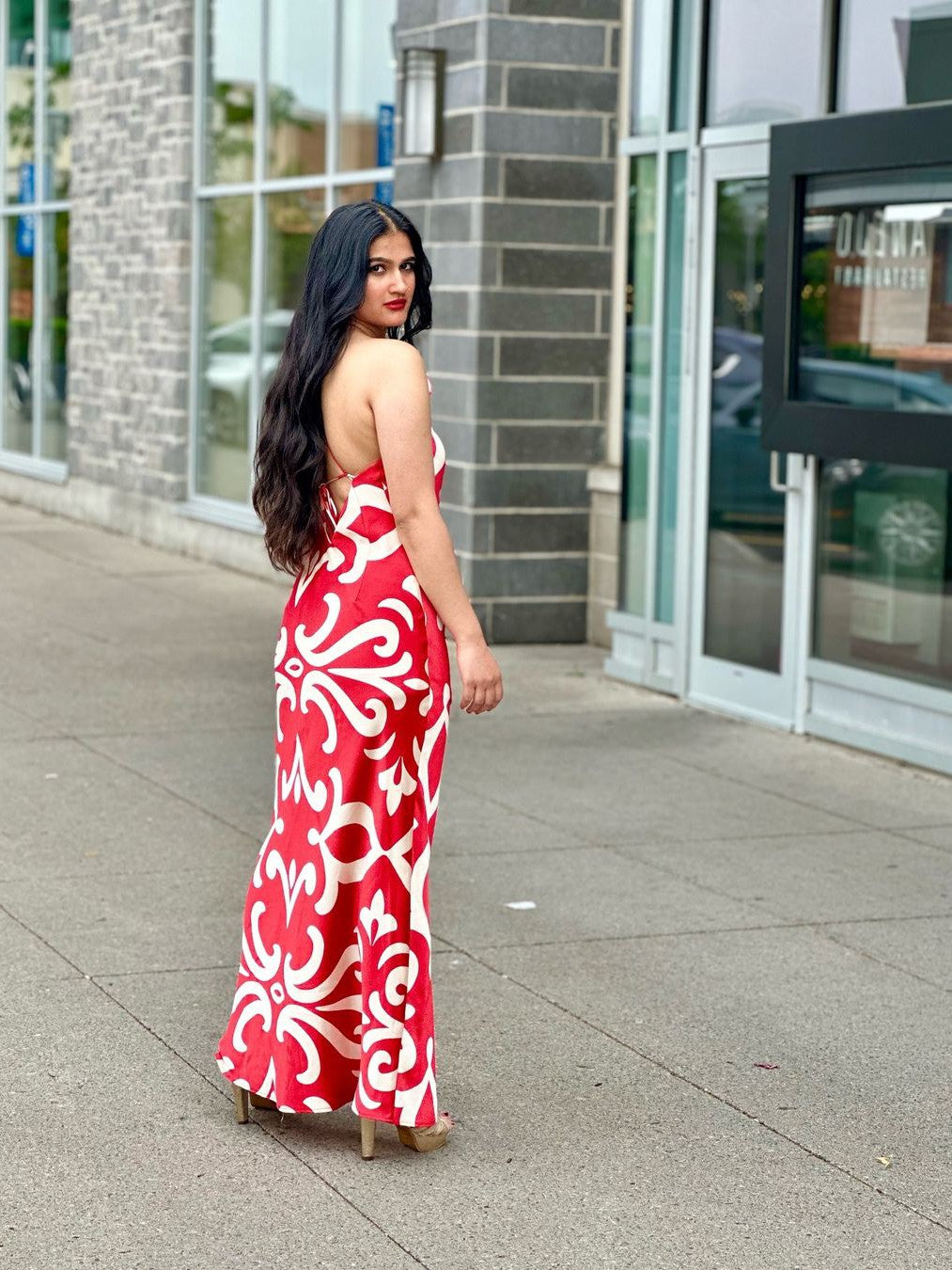 Woman in a red and white floral halter-neck backless maxi dress, standing on a sidewalk beside storefronts; back tied.