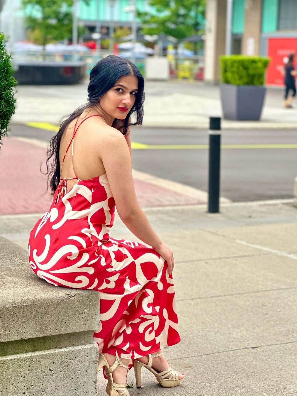 Woman seated on a concrete ledge in a red and white halter-back maxi dress with bold floral applique; beige heels.
