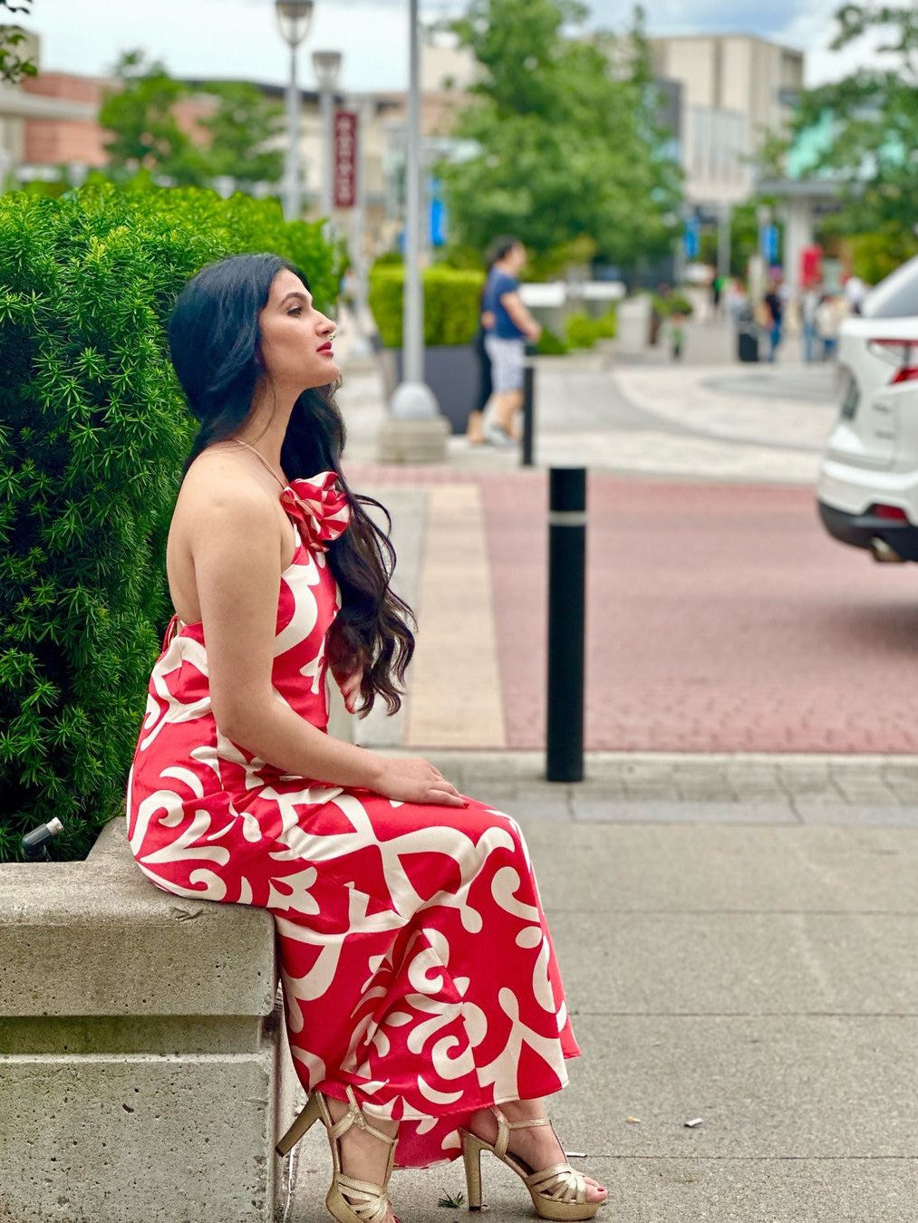 Woman in red and white floral halter-backless maxi dress seated on a concrete ledge beside green hedge in an urban street.