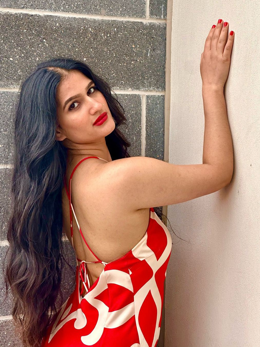 Woman in a red-and-white backless halter maxi dress, long dark hair, red lips, leaning on a beige wall.