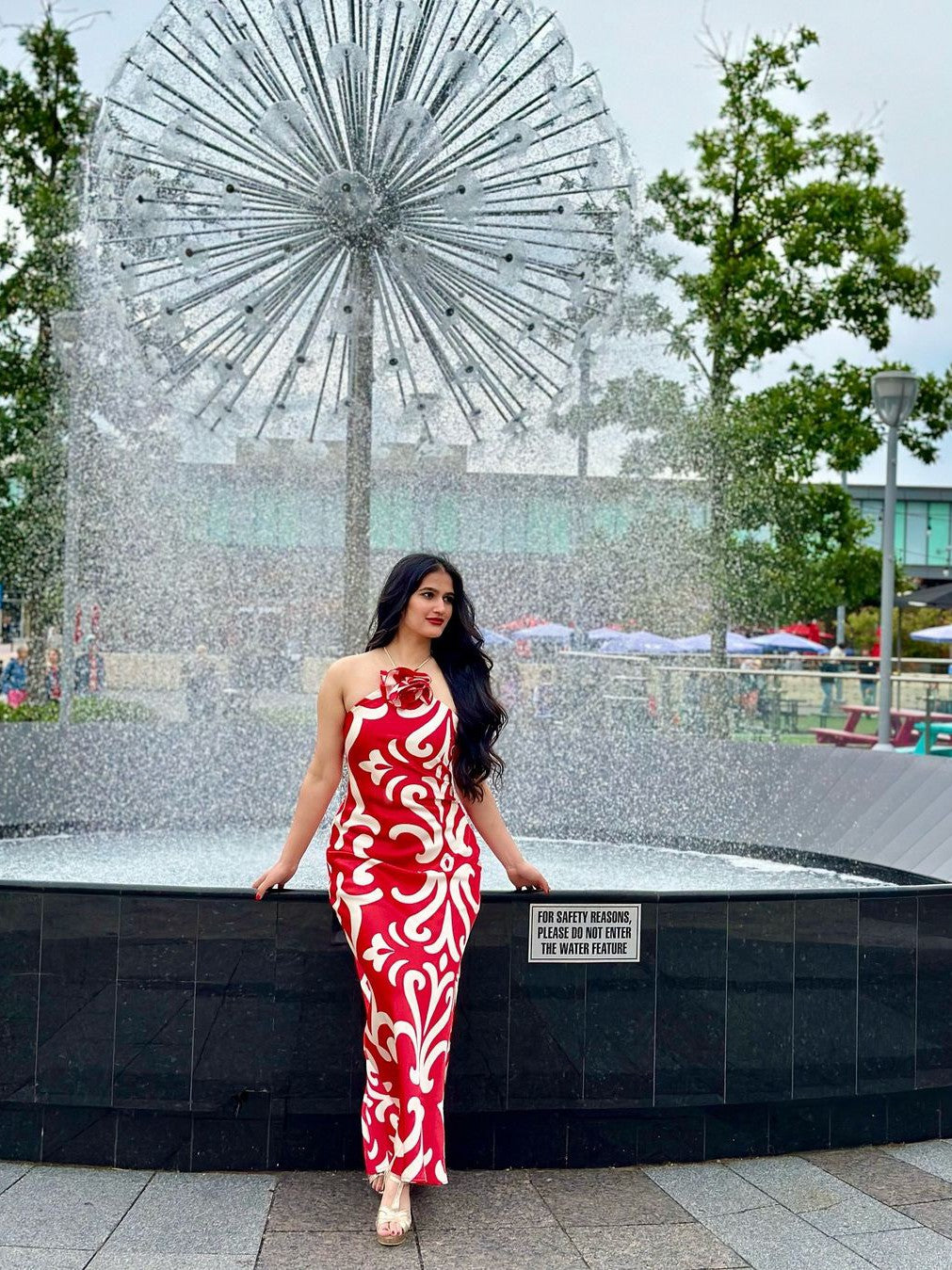 Woman in a red and white floral halter-neck backless maxi dress stands beside a circular fountain with a starburst metal sculpture behind.