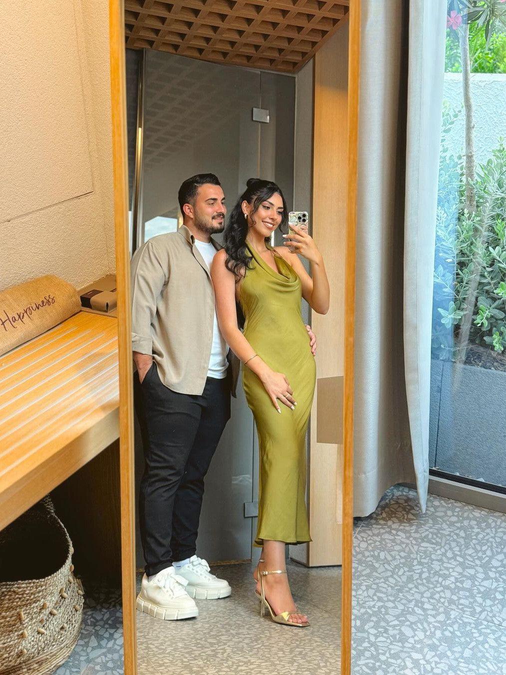 Couple in mirror selfie: woman in olive-green satin cowl-neck backless halter maxi dress with gold strappy heels; man in beige shirt.