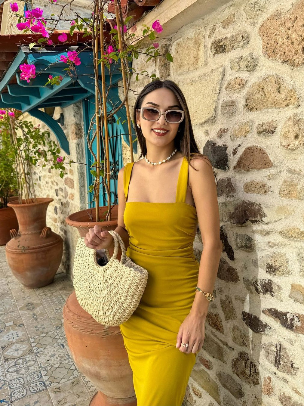 Woman in mustard yellow spaghetti-strap fitted dress beside a stone wall, carrying a woven straw bag.