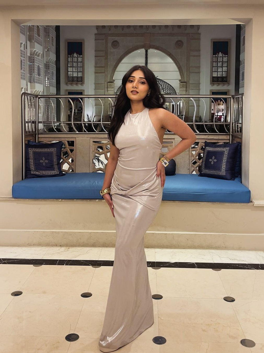 Woman in metallic champagne halter-neck backless ruched maxi dress, posing with hand on hip in a marble lobby with a blue cushioned bench.