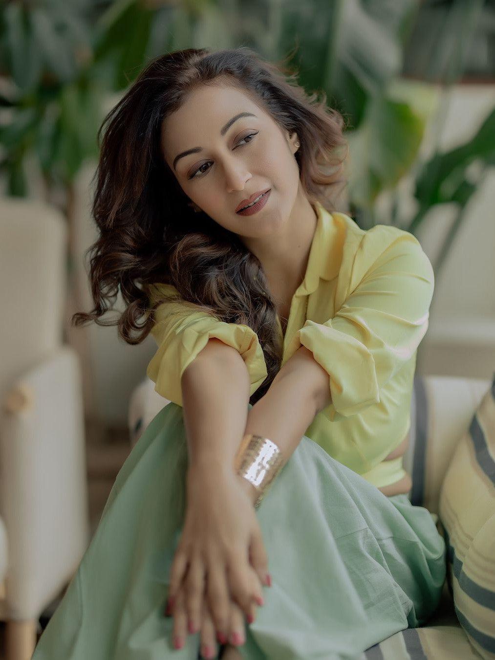 Woman in pastel yellow cropped blouse and mint green linen pants, seated on a sofa with a gold cuff bracelet.