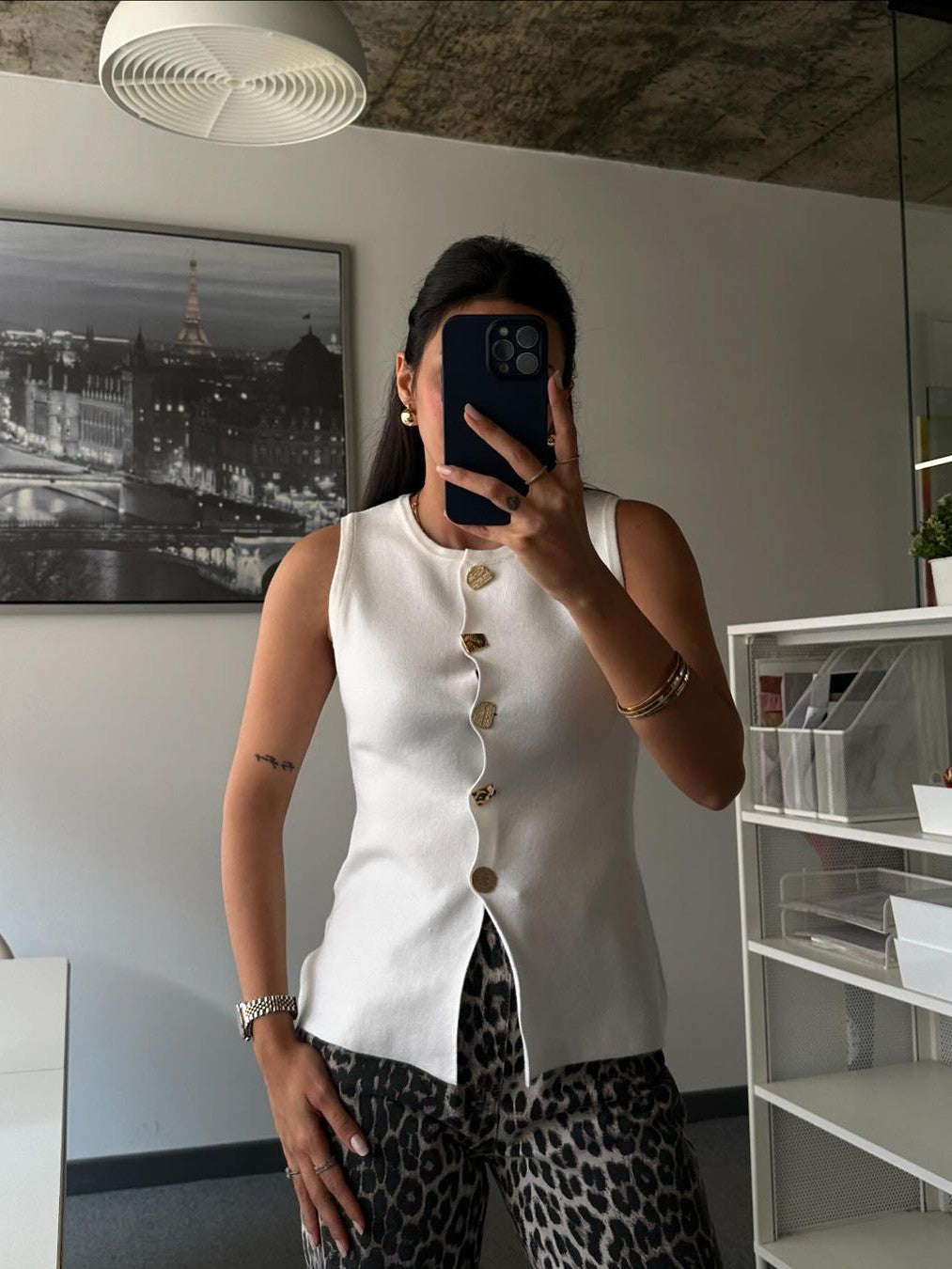 White sleeveless vest with gold buttons and curved front placket worn by a woman indoors, leopard print pants.