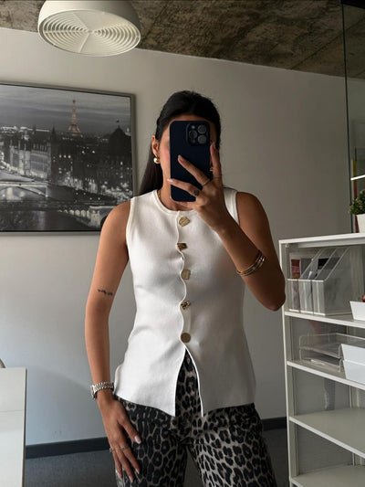 White sleeveless vest with gold buttons and curved front placket worn by a woman indoors, leopard print pants.