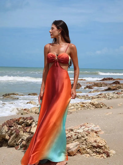 Rocky beach scene with a woman wearing a gradient orange-to-teal tie-dye cutout maxi dress, thin straps, and front knot.