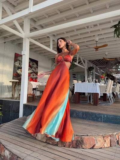 Woman in gradient tie-dye cutout maxi dress with twisted bust, orange to teal hues, on a wooden deck at a restaurant.