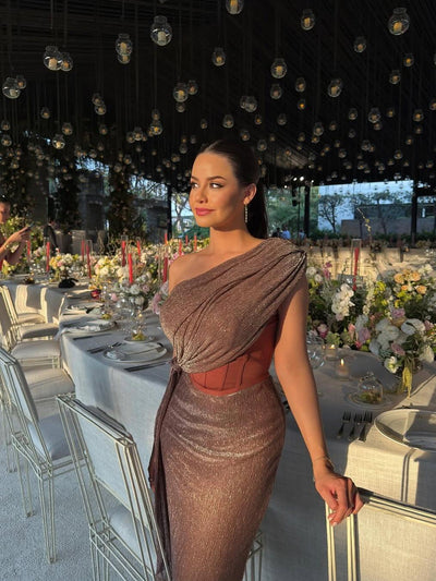 Woman in a shimmery rose-gold one-shoulder draped corset dress with a cinched waist, posing beside a long, flower-filled banquet table.