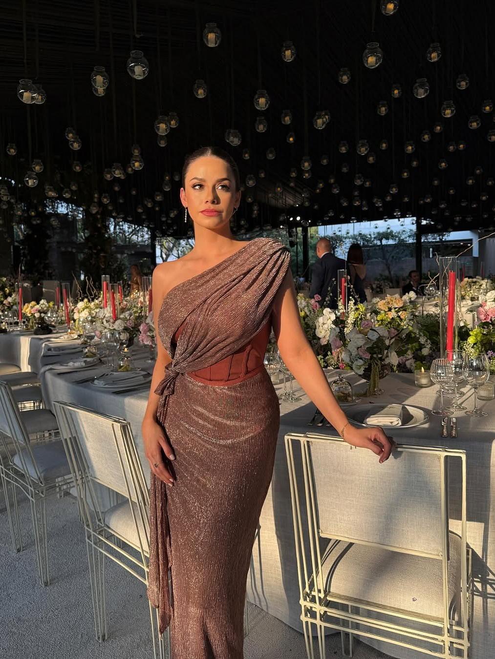 Woman in a rose-gold, shimmery draped one-shoulder corset dress with a wrapped bodice and glittery skirt at a formal banquet.